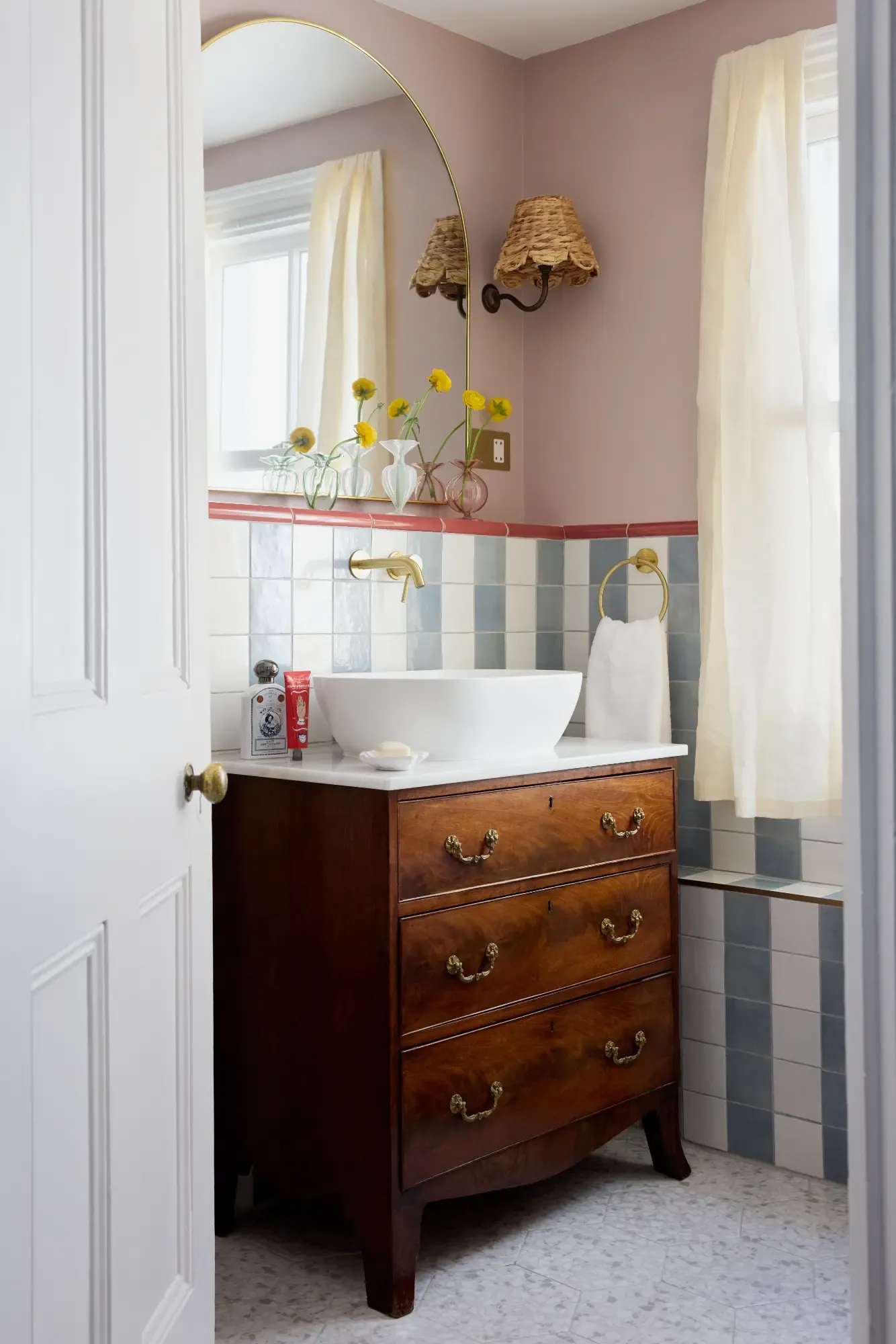 A white vessel basin on an antique mahogany chest of drawers, below an arched brass mirror and a wicker shaded wall light, against blue and white checkerboard tiling.