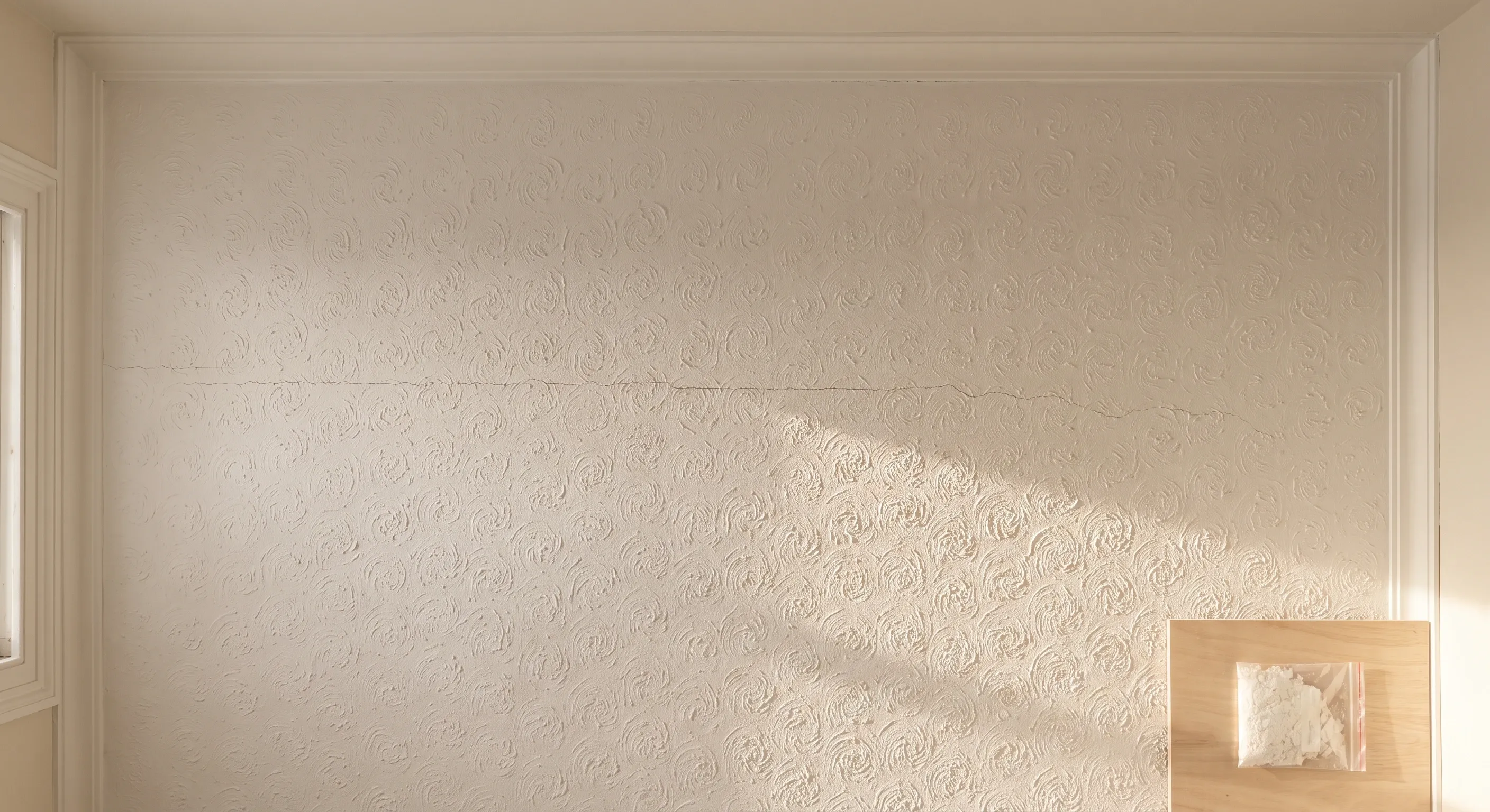 A stippled Artex ceiling seen from directly below in raking daylight, with a small sealed sample bag on a clean board at the edge of frame.