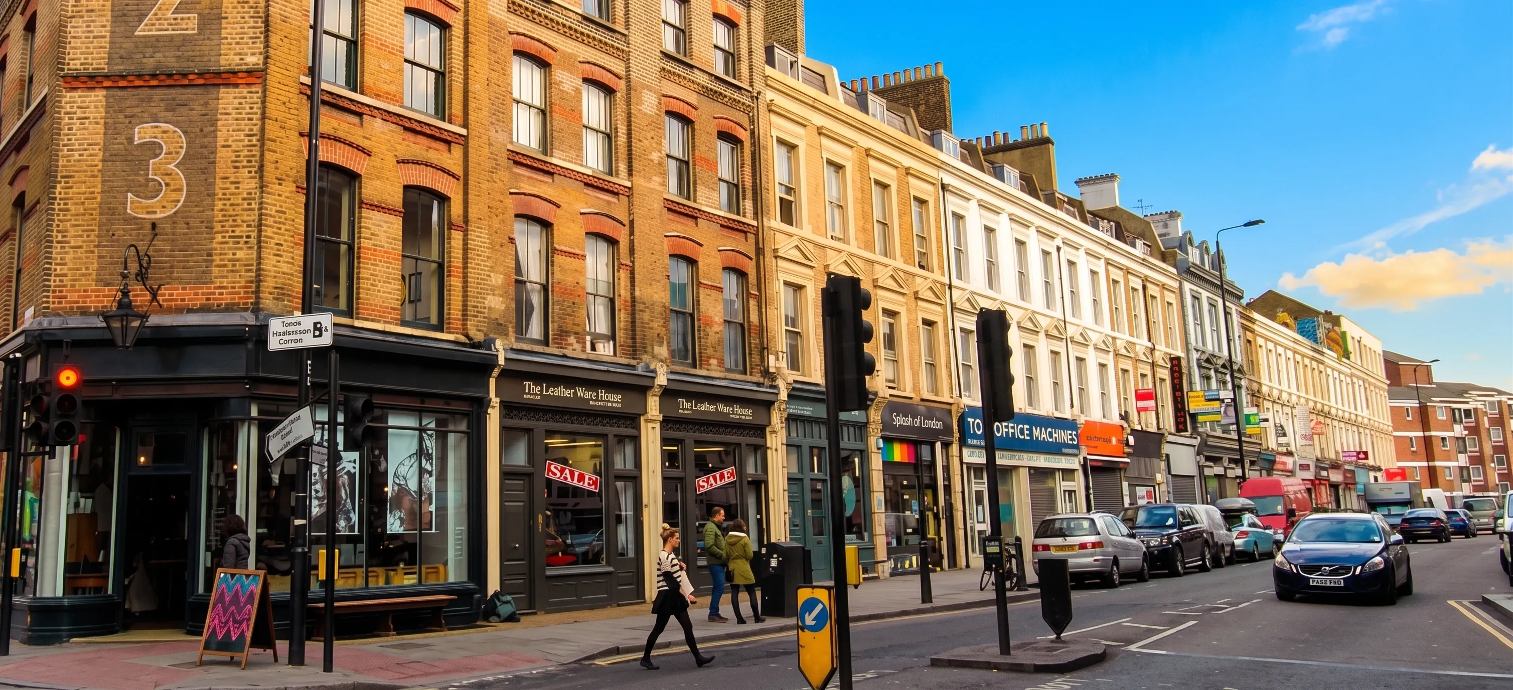 A row of Victorian brick buildings with shopfronts along a sunlit high street, with pedestrians on the pavement and traffic lights at the corner.