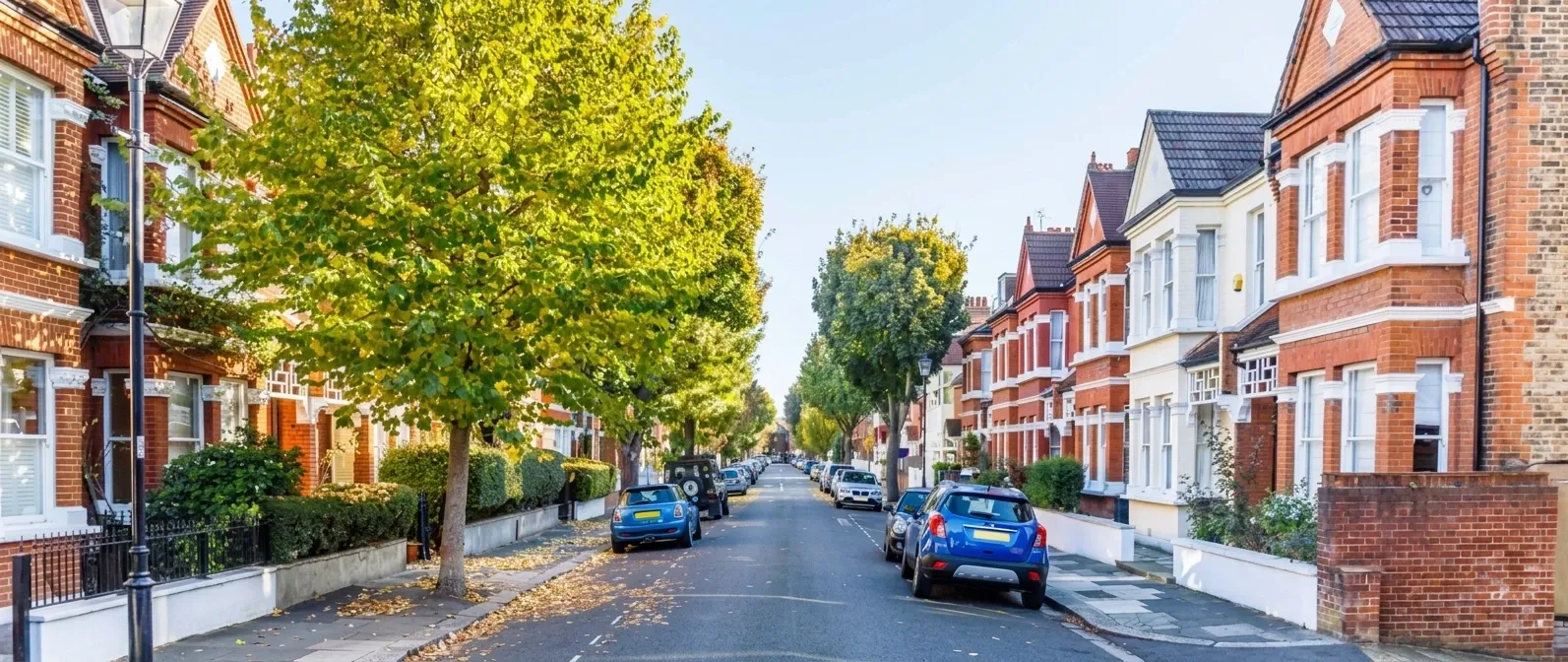 A tree-lined street of red brick Victorian terraces in Chiswick, with bay windows, autumn leaves on the pavement and cars parked along both sides.