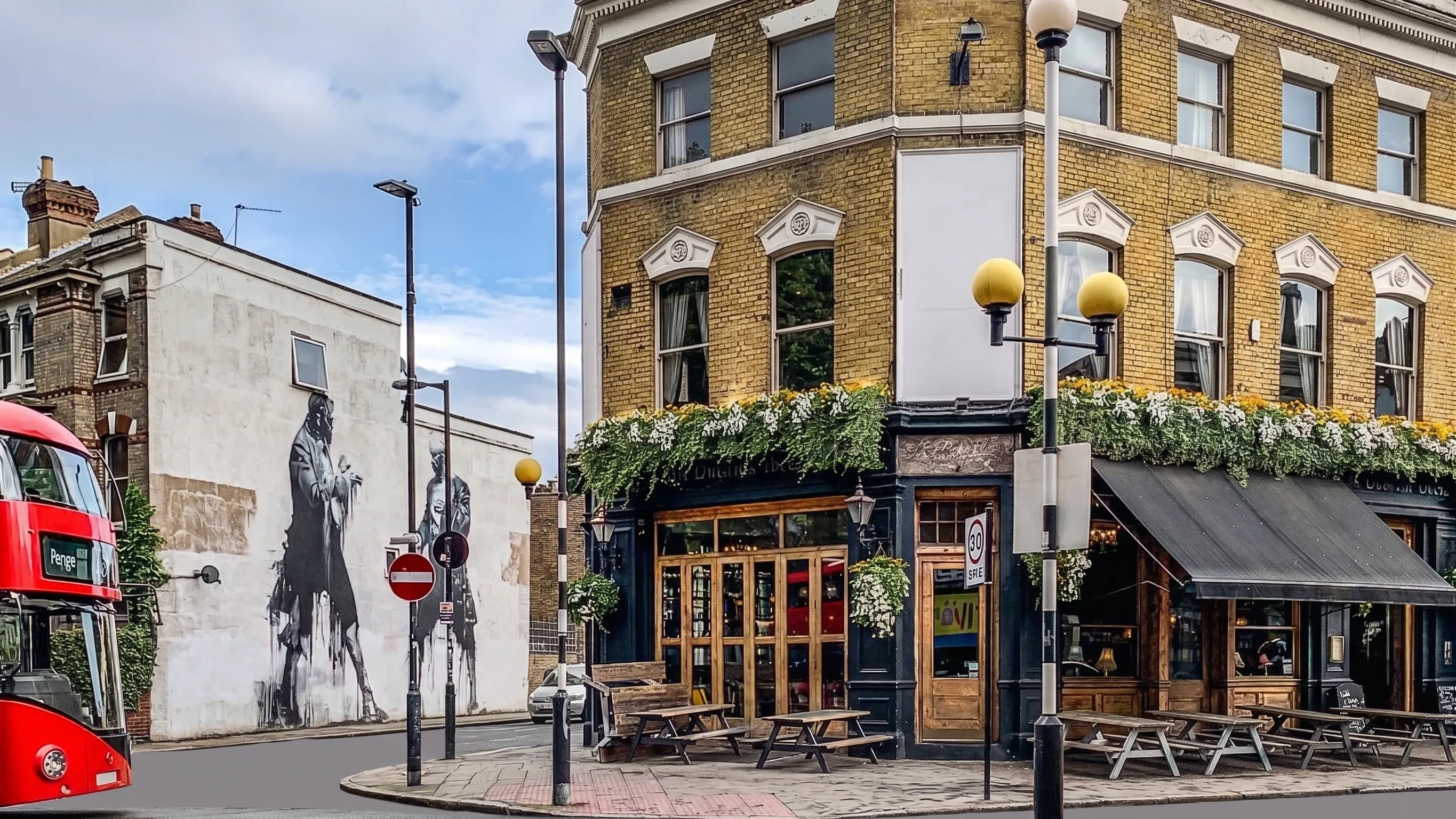 A corner pub in yellow stock brick with window boxes and picnic benches in East Dulwich, beside a large monochrome mural on a neighbouring gable end.