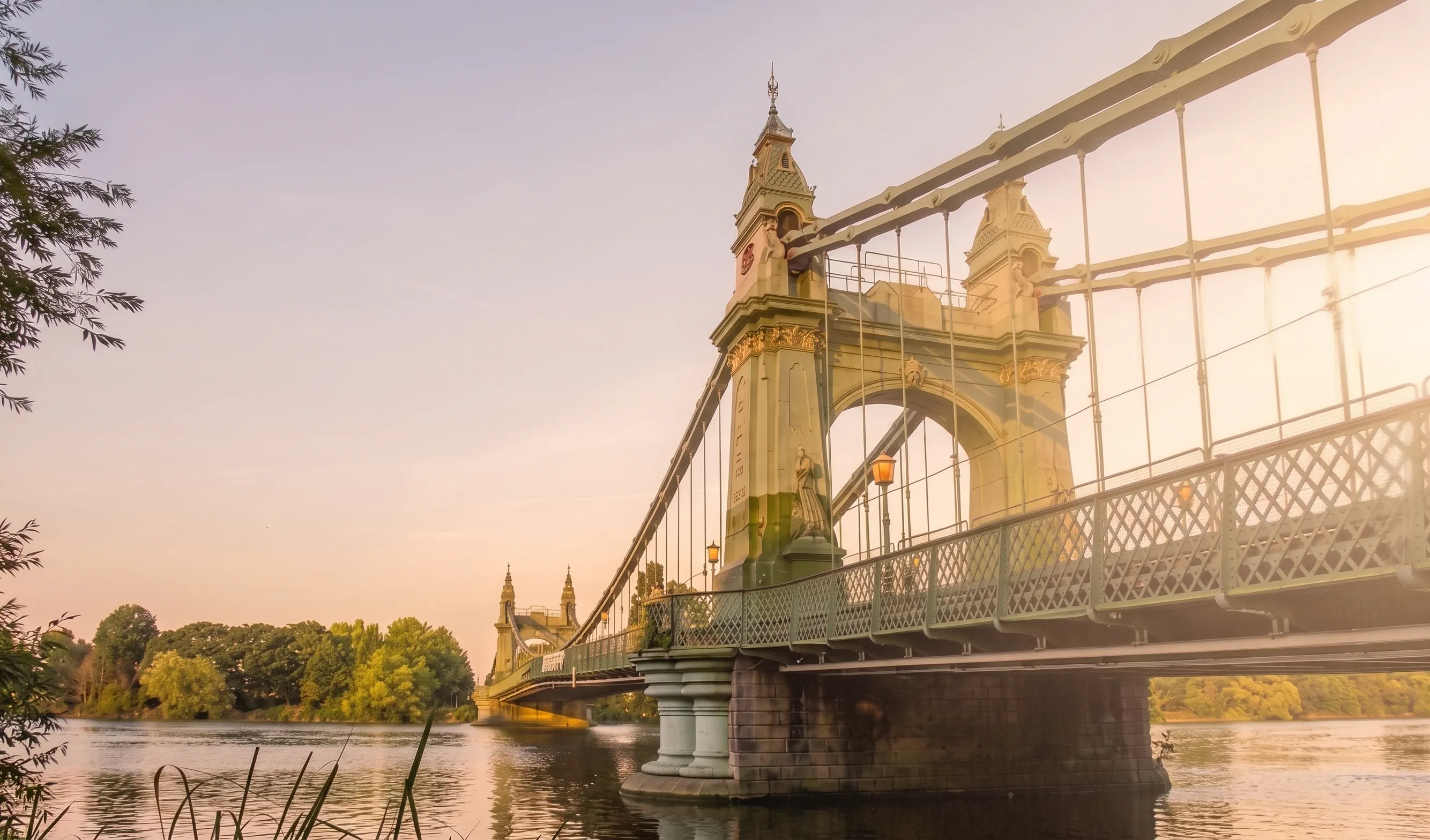 Hammersmith Bridge's green and gold suspension towers seen from the riverbank at sunset, with the Thames and trees on the far bank.