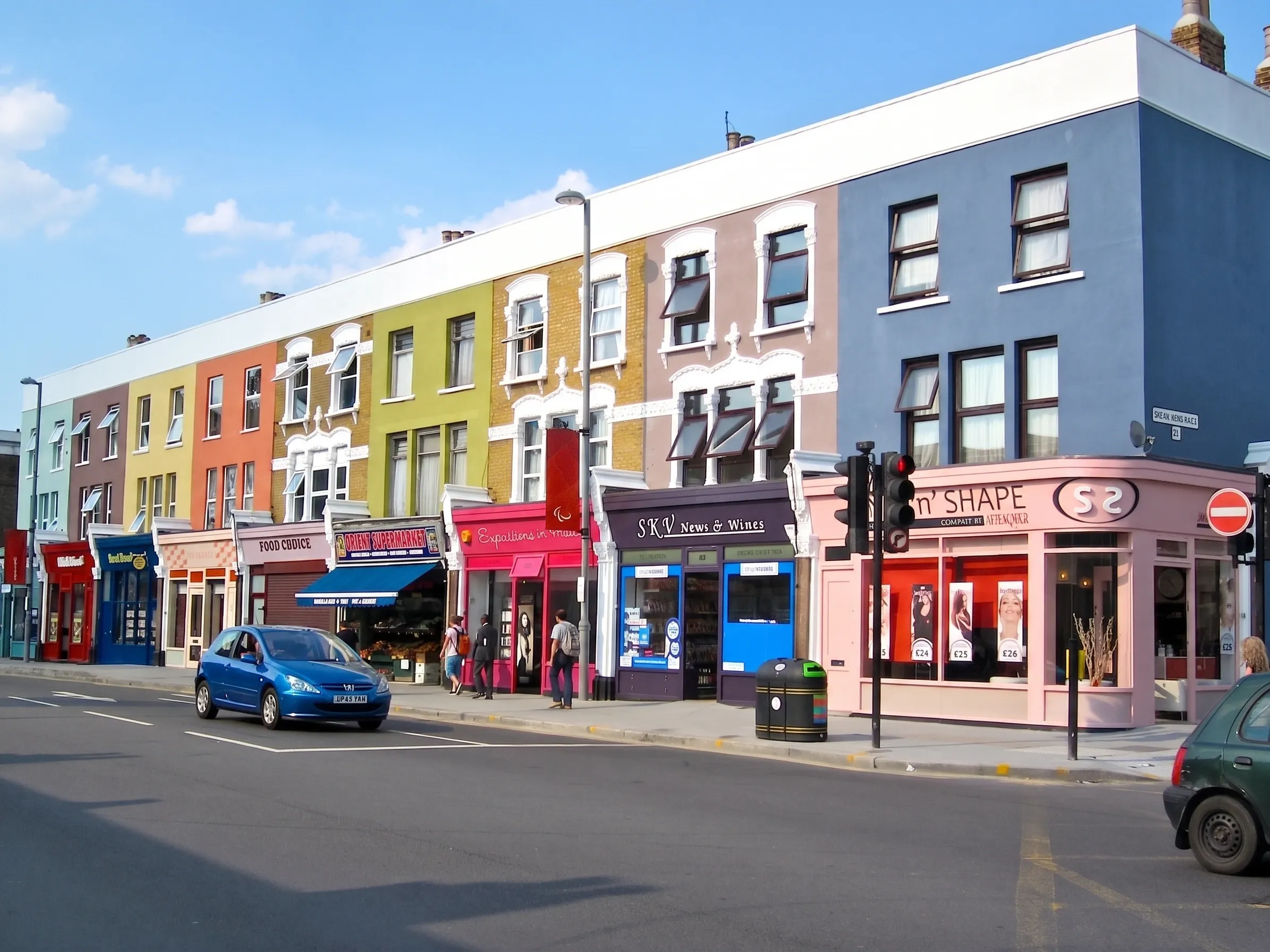 A terrace of brightly painted shopfronts on a parade in Leyton, with a supermarket, newsagent and salon at street level and flats above.