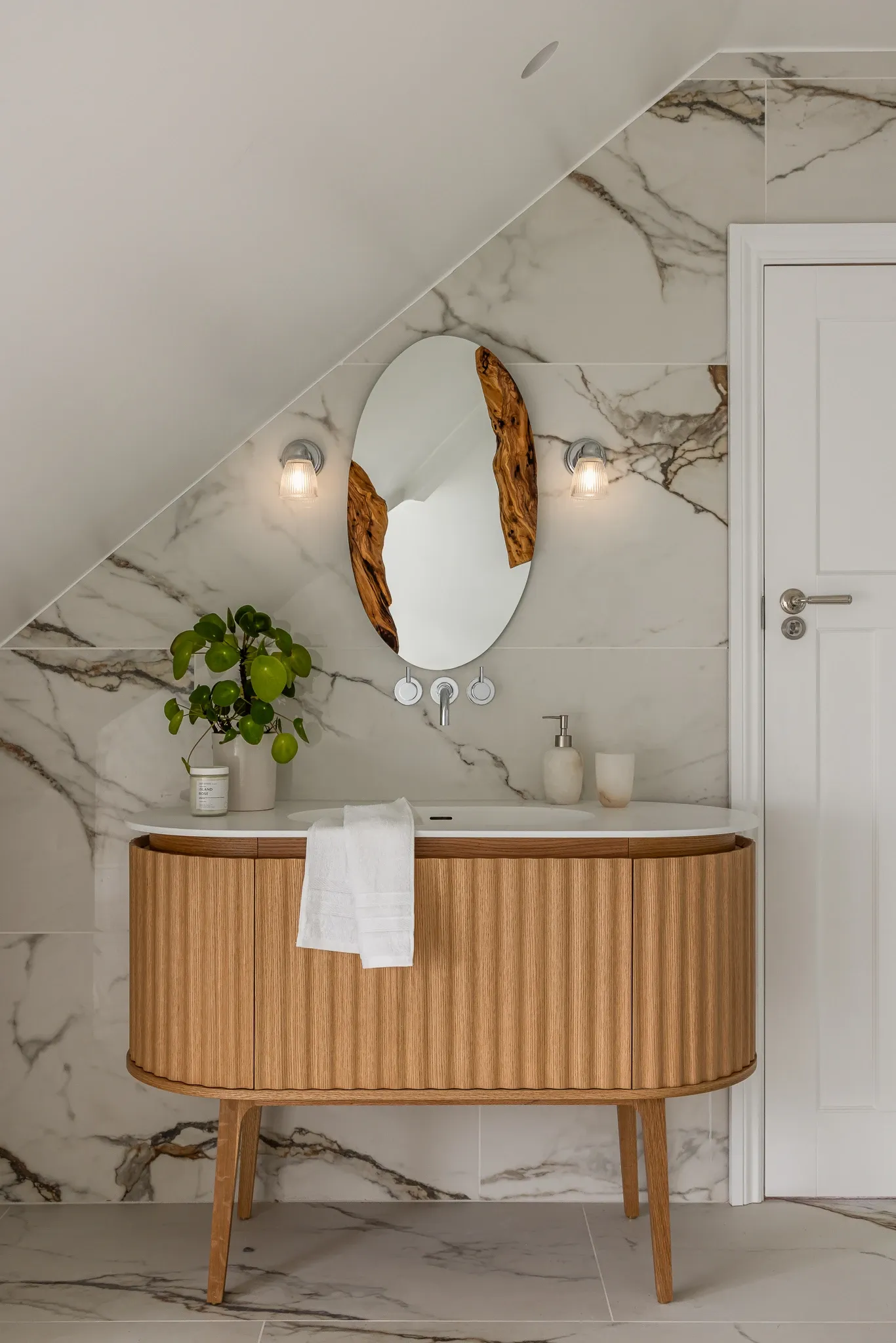 Fluted oak vanity on tapered legs beneath an oval mirror inset with olive wood, set against marble tiling under a sloped loft ceiling.