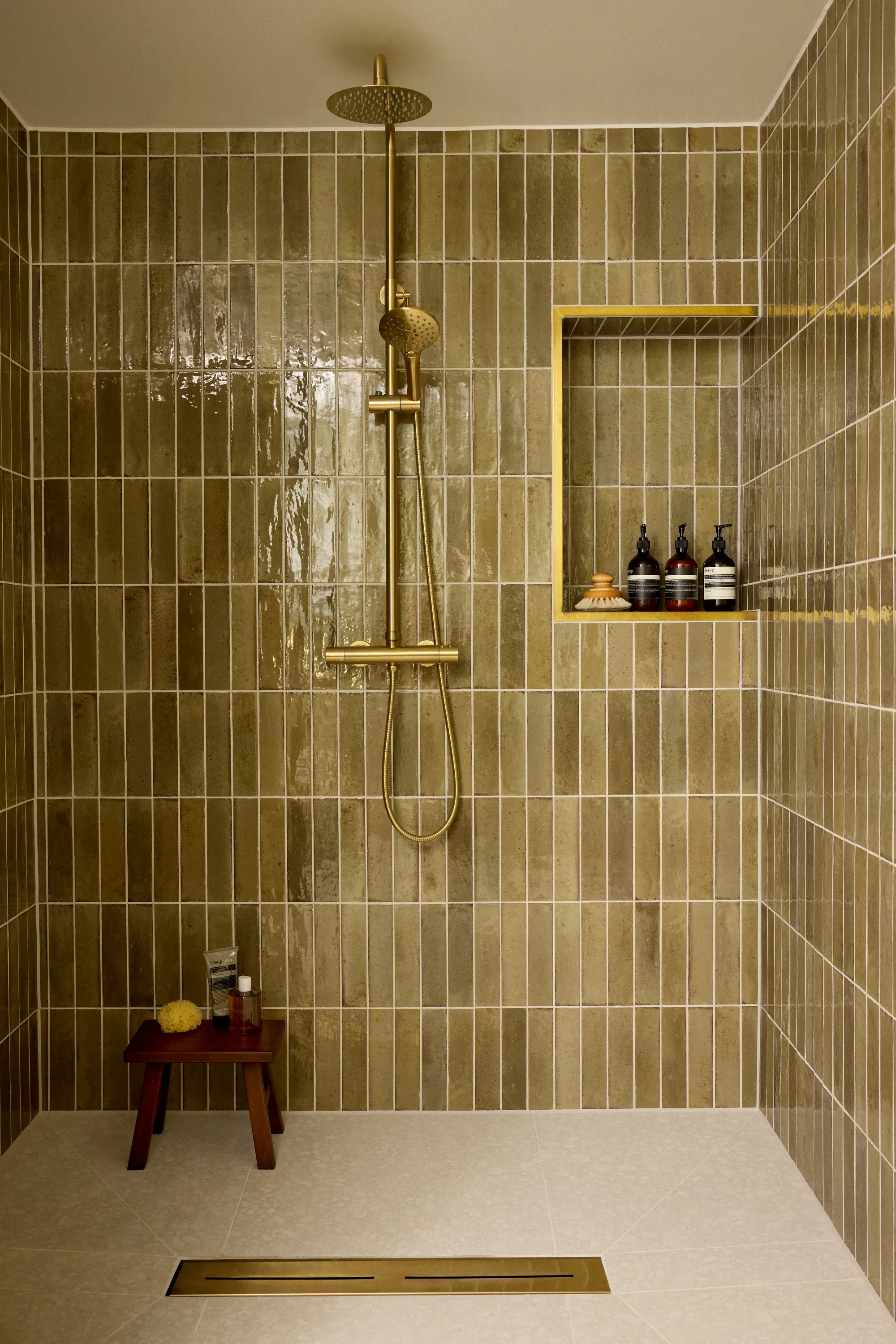 A walk-in shower tiled in glossy olive green stacked tiles, with a brass riser shower, a brass trimmed niche and a teak stool on the shower floor.