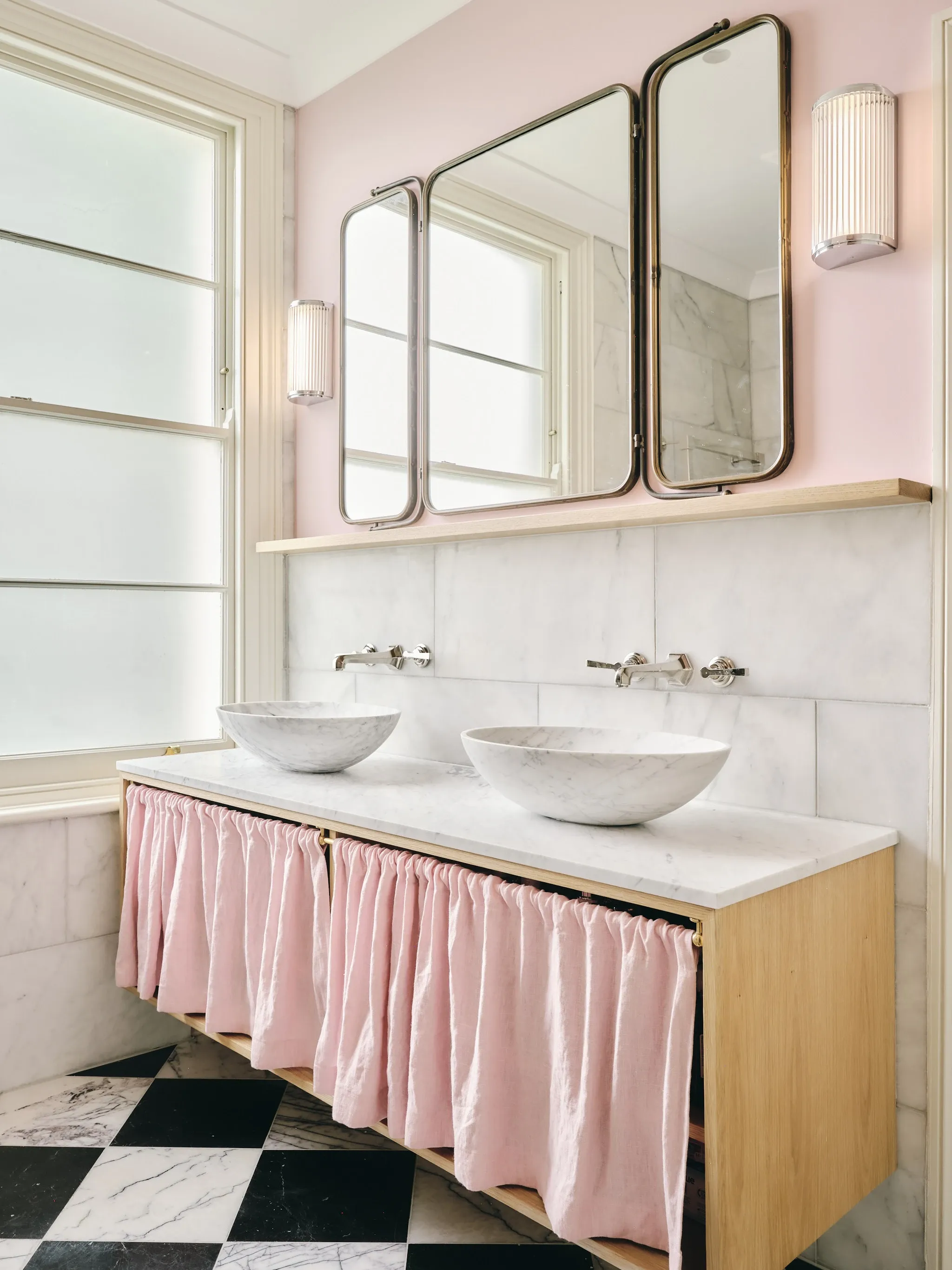 Two carved marble bowl basins on a marble top with a pink linen curtain beneath, in front of three hinged brass mirrors above a black and white checkerboard floor.