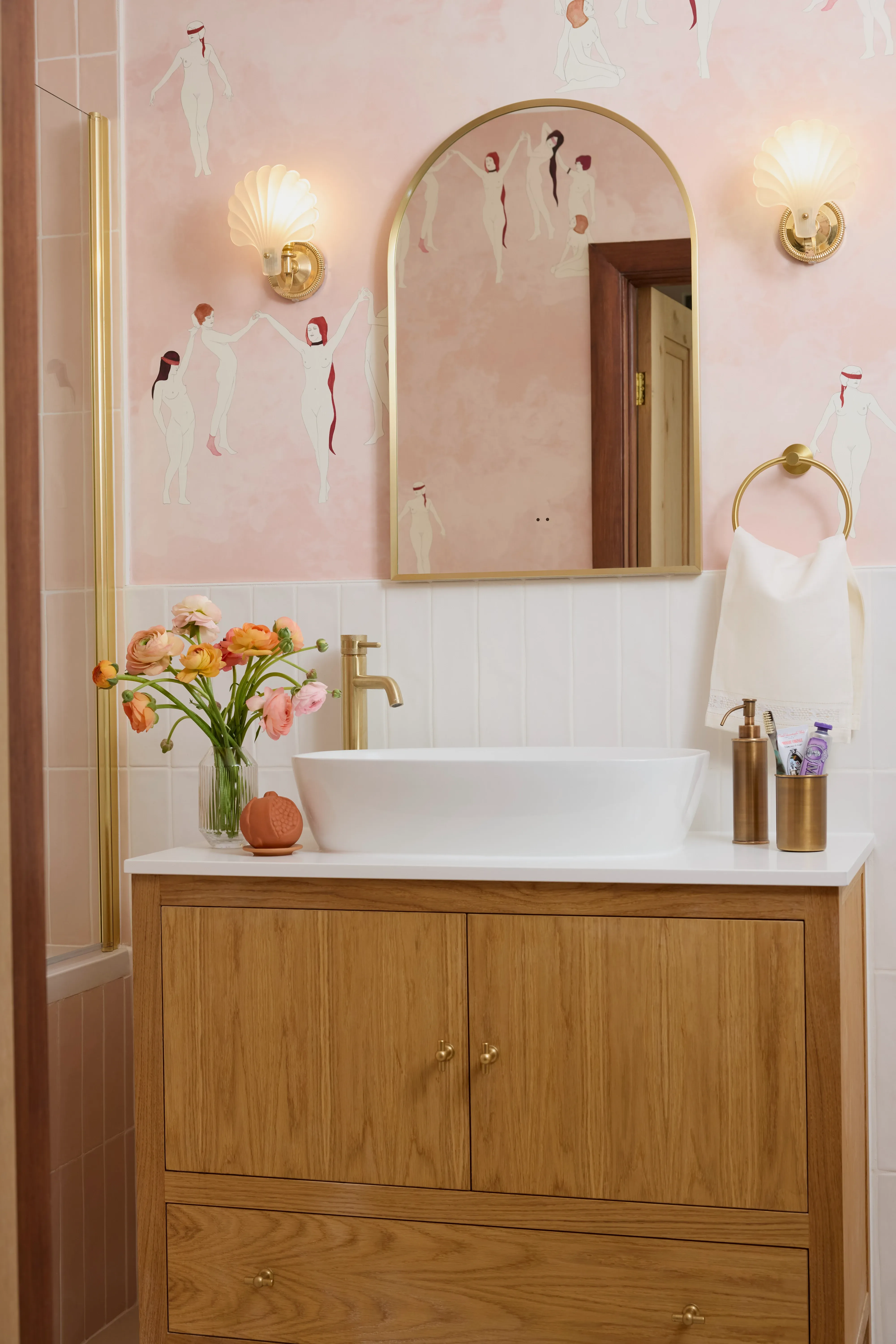 Oak vanity with a white vessel basin and brass tap below an arched brass mirror, against a pale pink wall painted with dancing figures.